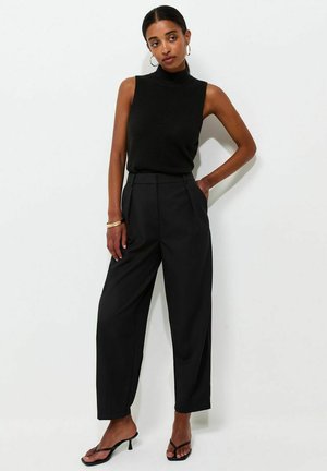 Femme portant un haut noir sans manches à col roulé, un pantalon noir large taille haute, des sandales noires à talons et des boucles d'oreilles et bracelets anneaux dorés.