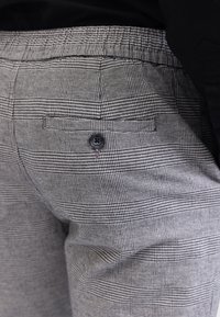 Gros plan sur un pantalon à carreaux gris avec une poche arrière noire boutonnée, porté avec un haut noir.