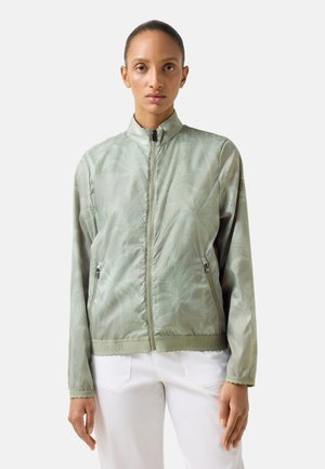 Woman wearing a light green zip-up jacket with high collar and two zipped pockets, paired with white pants, standing against a plain background.