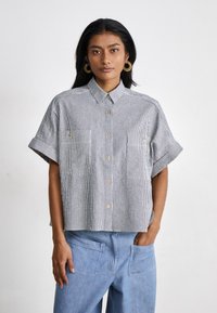 Camicia a maniche corte con colletto, a righe verticali blu e bianche, dotata di due tasche frontali e bottoni dorati. Con vestibilità rilassata.