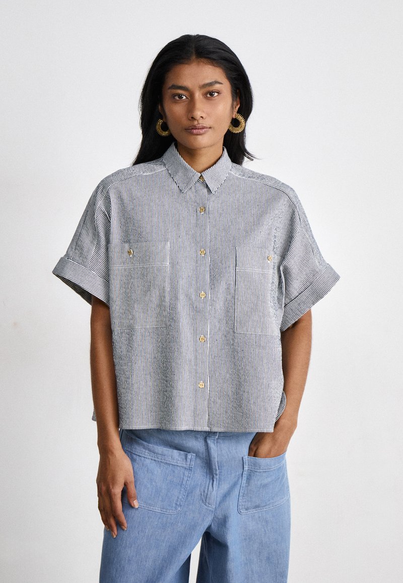 Camicia a maniche corte con colletto, a righe verticali blu e bianche, dotata di due tasche frontali e bottoni dorati. Con vestibilità rilassata.
