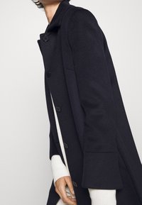 Dark wool coat with a structured collar, featuring six black buttons and loose sleeves. White sweater visible underneath.