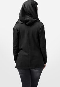 Personne portant une veste noire à capuche avec la capuche relevée, associée à un jean gris foncé, debout dos au spectateur.