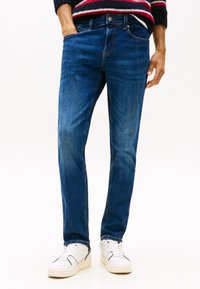 Modre kavbojke iz jeansa z ravnim krojem, petimi žepi in vidnimi šivi. Kombinirane z belimi supergami na nevtralnem ozadju.