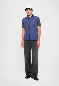 Colete acolchoado azul-marinho com gola alta, fecho zíper e detalhe do logotipo, combinado com uma camisa cinza de mangas curtas e calças de ganga escuras.