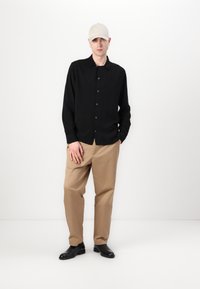 Schwarzes Langarmhemd, geknöpft, kombiniert mit beigen Hosen und schwarzen Schuhen. Das Model trägt eine hellfarbige Kappe und steht vor einem einfarbigen Hintergrund.