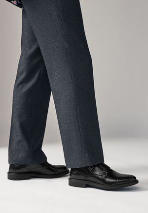 Piernas con pantalones de vestir a cuadros gris oscuro y zapatos de vestir de cuero negro con doble hebilla tipo monk sobre un suelo gris claro.