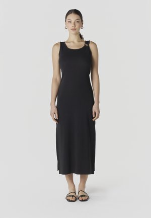 Ženska stoji v črni midi obleki brez rokavov z obročem na levi rami in črnimi sandalami z odkrito peto.