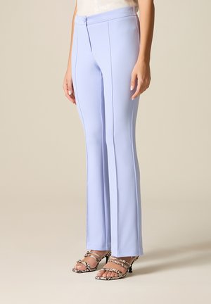 Persona che indossa pantaloni sartoriali azzurri chiaro con cuciture frontali e sandali con tacco alto in pelle di serpente, in piedi su sfondo beige.