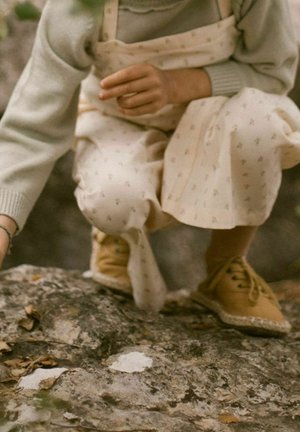Chaussures beiges à surface texturée et lacets, portées avec un pantalon ample de couleur claire orné d'un petit motif floral.