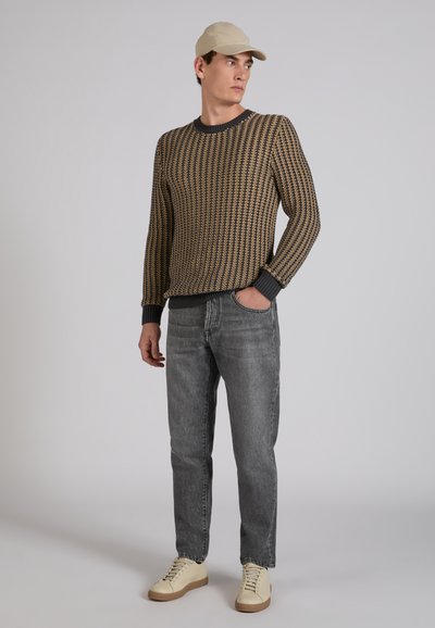 Hombre con gorra beige, suéter marrón y negro con patrón, jeans grises y zapatillas crema, de pie con una mano en el bolsillo, mirando hacia un lado.