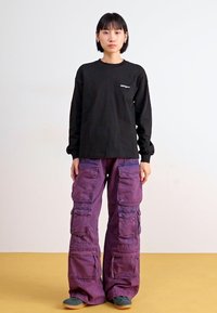 Chemise noire à manches longues associée à un pantalon cargo violet ample avec plusieurs poches et une finition texturée. Des chaussures grises complètent la tenue.