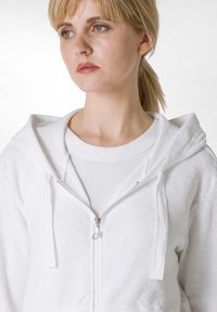 Weiße Zip-Hoodie aus einem weichen, strukturierten Stoff; mit Kapuze, Kordelzug und glatt zuziehbarem Reißverschluss. Über einem weißen, gerippten T-Shirt getragen.