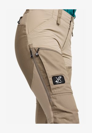Beige Outdoor-Hosen mit mehreren Reißverschlusstaschen, verstärkten Nähten und einem schwarzen RVRC-Logo-Patch am Oberschenkel. Glattes, strapazierfähiges Material.