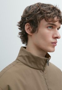 Brun jakke med lynlås og høj krave, glat tekstur og diskret glans. Har en lille sølvfarvet lynlås og minimalistisk design.