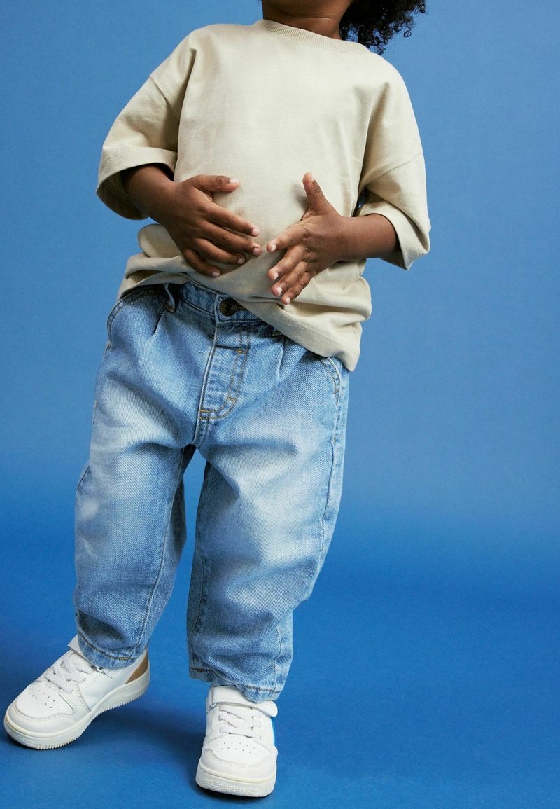 Enfant portant un t-shirt beige, un jean bleu clair ample et des baskets blanches, debout devant un fond bleu, mains près du ventre.