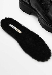 Mustat tekoturkisvuoret, joissa on pehmeä rakenne ja pyöreä muoto, varustettuna tarralla, jossa lukee "Massimo Dutti." Aseteltu mustan saappaan viereen.