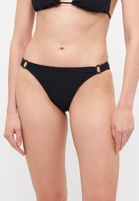 Black bikini bottom with a textured diamond pattern, featuring circular hardware at the sides and a low-rise cut. Soft fabric.