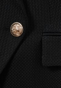 Nahaufnahme eines schwarzen, strukturierten Blazerstoffs mit einem goldenen, dekorativen Knopf mit erhabenem Löwendesign und einer Paspeltasche mit Klappe.