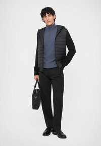 Casaco puffer preto com fecho e capuz, sobreposto a um suéter azul-marinho com fecho de zip e calças pretas. A segurar uma mala preta.