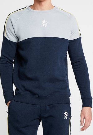 Homme portant un sweat-shirt bleu marine et gris clair ainsi qu'un pantalon assorti avec un logo blanc et des bandes jaunes sur les épaules et les côtés du pantalon.