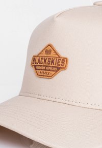 Gorra beige con un diseño estructurado, que presenta una tela texturizada y un parche de cuero marrón que muestra "BLACKSKIES PREMIUM SUPPLIES MMIX".