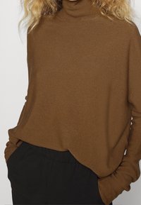 Brązowy sweter z golfem wykonany z miękkiego materiału, o luźnym kroju i długich rękawach, zestawiony z czarnymi spodniami.