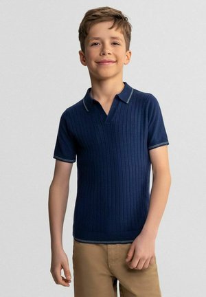 Chłopiec stojący i uśmiechający się, ubrany w granatową, prążkowaną koszulkę polo z krótkim rękawem oraz khaki spodnie na jednolitym tle.