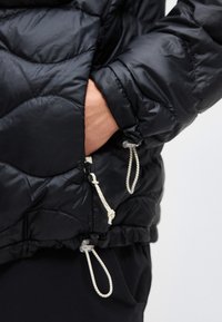 Mână în buzunarul unei jachete negre matlasate, cu șnururi albe reglabile pe mânecă și la tiv.