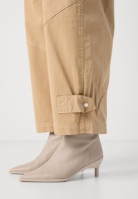 Beige, leveled pants dengan jahitan dan detail kancing snap, dipadukan dengan sepatu bot pergelangan kaki kulit beige dengan ujung runcing dan heel kecil.