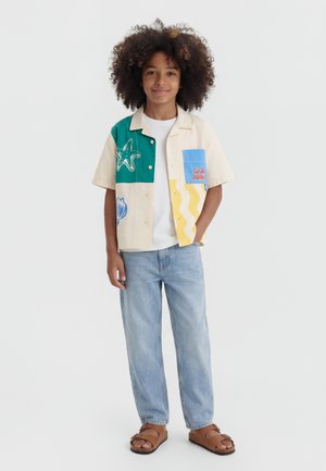 Kind mit lockigem Haar, das ein buntes kurzärmeliges Hemd mit Knöpfen, hellblaue Jeans und braune Sandalen trägt, vor einem schlichten Hintergrund stehend.