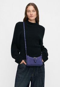 Piccolo borsa a tracolla in suede viola con dettagli in fibbia argentata e tracolla regolabile, esposta su un maglione nero a coste e jeans.