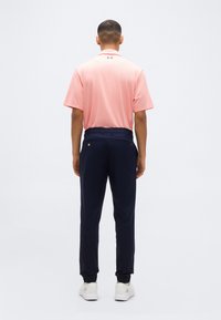 Άνδρας που στέκεται με την πλάτη προς τα εμπρός, φοράει ροζ πουκάμισο πόλο, σκούρο μπλε παντελόνι και λευκά αθλητικά παπούτσια, μπροστά σε απλό φωτεινό φόντο.