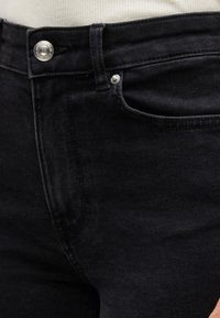 Μαύρα τζιν από τζιν ύφασμα με κουμπί και ραφές, φορεμένα πάνω από λευκό ριμπ ύφασμα τοπ.