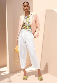 Lichtroze blazer over een bloemenpatroon top, gecombineerd met witte broeken en groene gevlochten sandalen. Een gele tas maakt de look compleet.