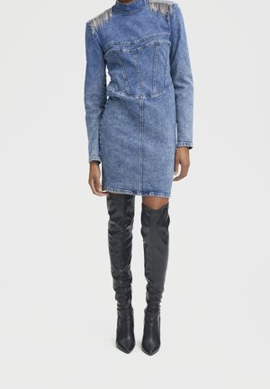 Person iført en blå denim minikjole med rhinestone frynser på skuldrene og sorte læder støvler over knæet med spids, stående.