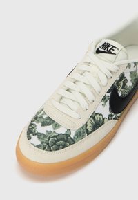Sneaker Nike bianca e beige con motivo floreale verde, logo swoosh nero, lacci bianchi e suola in gomma color caramello.