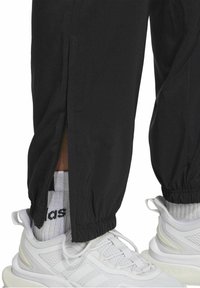 Schwarze, schmal zulaufende Jogginghosen mit Reißverschlüssen an den Knöcheln und elastischen Bündchen, kombiniert mit weißen Sportschuhen und gestreiften Socken. Glatte Stoffstruktur.