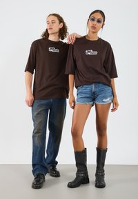 Camisetas oversized marrones con logo blanco, pantalones cortos de mezclilla azules y botas negras de rodilla con acentos de tachuelas. Tejido suave y ajuste casual.
