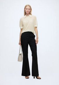 Femme portant un haut crème à manches courtes boutonné, un pantalon noir taille haute à jambes larges, des talons ouverts noirs et tenant un sac à main beige.