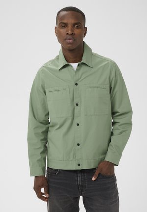 Mand iført lysegrøn knappet jakke med klaplommer, sorte jeans, stående foran en enkel lys baggrund.