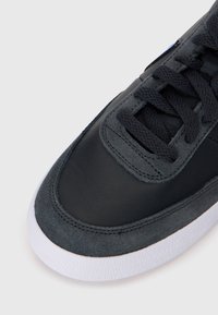 Dessus de chaussure en cuir noir et en daim avec une texture lisse, doté de lacets noirs, d'une semelle en caoutchouc blanc et de détails de couture renforcés.