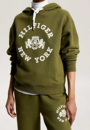 Osoba w oliwkowej zielonej bluzie z kapturem i spodniach dresowych z białym napisem "Hilfiger New York" oraz herbem na obu elementach.