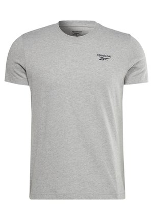 Mężczyzna w szarej koszulce Reebok, granatowych spodniach dresowych, białych trampkach, trzymający dużą niebiesko-czarną torbę sportową za uchwyty.