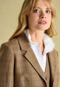 Hnedý károvaný blazer s ostrými klopami, detailným podšívaním a bielou košeľou s volánovým golierom viditeľnou. Mäkká textúra, štruktúrovaný dizajn.