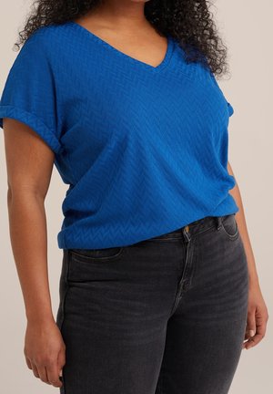 Nainen, joka pukeutunut lyhythihaiseen siniseen, teksturoituun V-kauluspaitaan ja korkeavyötäröisiin mustiin farkkuihin, seisoo yksinkertaisen taustan edessä.