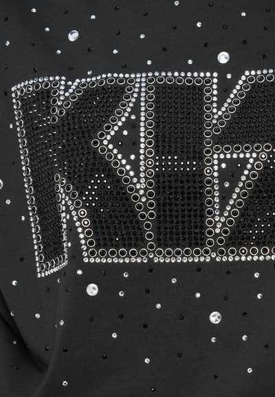 Black fabric featuring a large, studded logo densely embellished with clear and black rhinestones, accented by scattered crystal-like details.