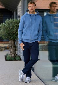 Felpa con zip blu e fodera grigia chiara, abbinata a pantaloni blu navy e sneakers grigie. L'ambientazione presenta muri in pietra e vegetazione.