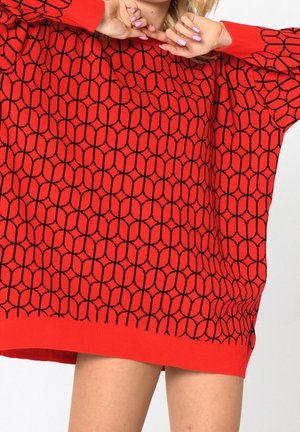 Frau trägt ein übergroßes rotes Pullover mit schwarzem, geometrischem sechseckigem Muster, posiert mit den Händen in der Nähe des Gesichts und sichtbaren Beinen unter dem Saum.
