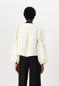 Blouse en satin crème avec manches bouffantes, col noir, détails plissés, design court, associée à un pantalon noir.